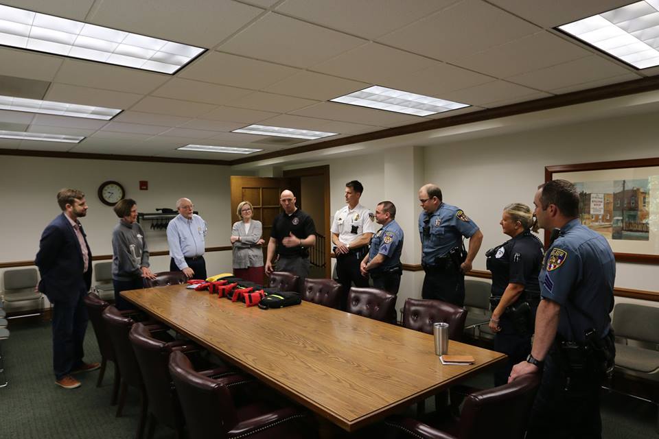 Webster Groves Police Department personnel and supporters stand around a table holding AED cases